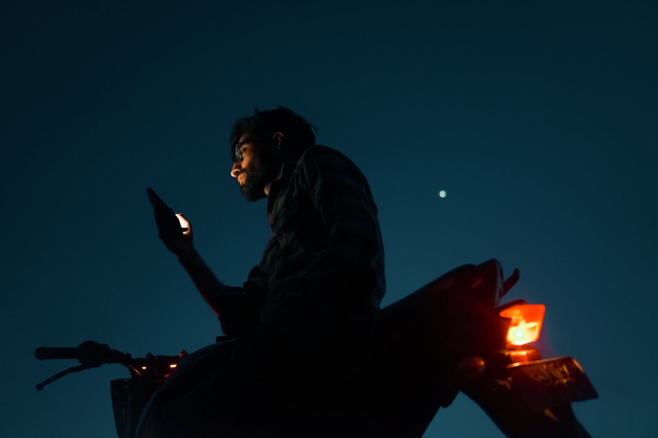 Silhouetted man on motorcycle using smartphone under night sky with star visible.